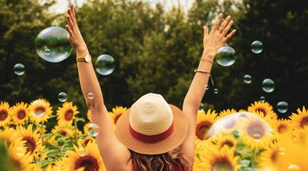 back of a woman wearing a hat with her hands up in a field of sunflowers